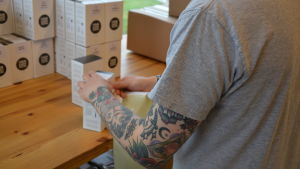 Största delen av arbetsdagen på Haahtis lilla kafferosteri går åt till att packa kaffe för kunderna. Rosteriet har ungefär 20 kunder i dag. De flesta finns i Finland, men en del av lådorna skickas utomlands. En person i grå t-skjorta viker kaffepaket.