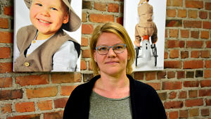 Virpi Hagström, verksamhetsledare Vasa mödra- och skyddshem Virpi Hagström står framför en tegelvägg prydd av tavlor med fotografier av barn.