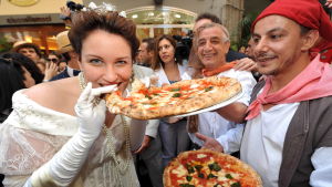 Savoijin kuningatar Margheritaksi pukeutunut nainen haukkaa nimeään kantavaa pizza margheritaa pizzakokkien ja yleisön ympäröimänä ko. pizzan 120-v-juhlassa Napolissa v. 2009.
