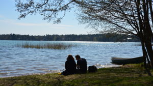 En kvinna och en man sitter vid strandbrynet en solig dag. Havet glittrar och man ser dem i motljus.