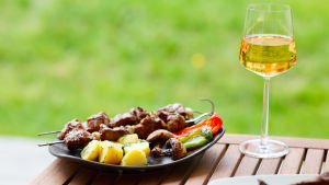 Hungriga getingar söker sig till mat och söta drycker i slutet av sommaren. En portion med grillmat står på ett utebord i trä. Ett glas med vitt vin står bredvid.