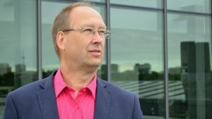 Jörgen Wiik. En man i kostym och skjorta utan slips blickar bort åt fjärran. I bakgrunden syns en reflektion av tak i fönster.