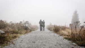Speciellt när det blir mörka, kalla tider är det svårare att ses utomhus och det blir tungt att vara isolerad. Två personer håller om varandra och går längs en grå väg. Bilden är tagen bakifrån.