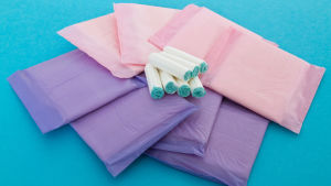 I Finland har mensskydd en mervärdesskatt på 24 procent. Böcker och läkemedel har runt tio procent. Bild: Yle Oanvända dambindor och tamponger i en hög. Turkosblå bakgrund.