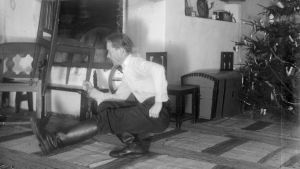 En man lyfter en stol med sin ena hand samtidigt som han är på huk med ena benet framåt. Bilden är tagen 1936 i Kulla, Strömfors.