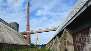 Sockerfabrikens skorsten. På bilden ser man skorstenen vid sockerfabriken på avstånd. Brevid den står en gul lyftkran. I förgrunden ser man två lagerbyggnader.