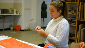 Elin Björkström är med och arrangerar stickgraffiti-workshopen i och med att hon är aktiv i slöjdlärararnas studentförening Arte på Åbo Akademi. Elin Björkström sitter och stickar i ett klassrum. Framför henne ligger mängder av garn i olika orange nyanser.