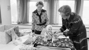 Moderskapsförpackningen 1977 presenteras. Moderskapsförpackningen 1977 presenteras av två kvinnor som klär på en docka.