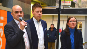 Stadsdirektören Mika Sivula (i mitten) samt sektorcheferna Pekka Puistosalo och Katri Kalske är de högsta tjänstemännen i Lojo. Två män och en kvinna i medelåldern står i ett köpcentrum. Männen har mörk kavaj och vit skjort, kvinnan mörk kavaja och blå tröja. Mannen längst till vänster håller en mikrofon i handen.