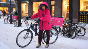 Många av restaurangmatsbuden använder egen cykel i jobbet, oberoende av årstid. Bild på Anton Salmenkylä i rosa jacka och hans cykel med en rosa termobox bakpå.