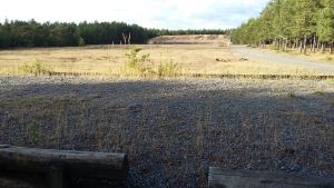En av de skjutbanor i Syndalen där grundvattnet ska skyddas bättre. En skjutbana på sandig mark. I förgrunden två stockar, i bakgruden syns en vall, på sidorna skog. Sol och skugga.