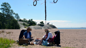 I år ordnas det picknick intill Casino i Hangö igen. Ett par har picknick på en strand. I bakgrunden syns en midsommarstång.