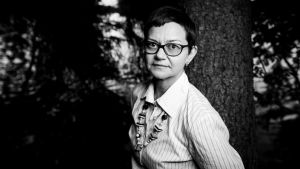 Nainen, Anna-Maija Halonen nojaa puuhun ja katsoo kameraan.