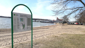 Infotavlor informerar om vattenkvaliteten. En skylt vid en strand där det står Knipans badstrand. Till höger syns ett träd och bakom det några byggnader.