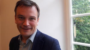 Gustav Hoffstedt säger att förändringen är välkommen - trots en ny skatt är det bra med regler på marknaden. Gustav tittar i kamerna, småler, invid ett fönster, träd i bakgrunden.