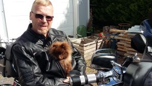 Thomas Wikholm iklädd svart läderjacka. Sitter på en motorcykel. Innanför läderjackan ser man hunden Demi, en liten brun hund av rasen pomeranian.
