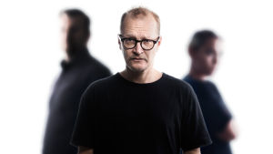 Kaapattu mieli -kuunnelman Petri (Janne Reinikainen) Janne Reinikainen katsoo etualalla kameraan, taka-alalla kaksi ihmishahmoa.