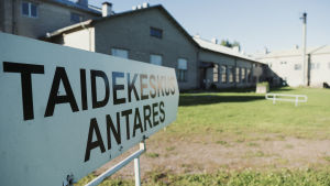 Näyttelyssä kiertelyn vastapainoksi laajalla pihamaalla on tilaa oleskella ja vaikka lasten temmeltää. Kyltti jossa teksti Taidekeskus Antares, taustalla taidekeskuksen rakennus.