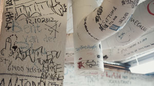 Tussilla kirjoitettuihin teksteihin ja graffiteihin peitettyjä valkoisia seiniä ja ilmastointiputkia.