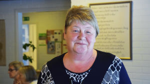 Mona Andersson. En kvinna ler på en bild, i bakgrunden syns en tavla och några personer i profil. Kvinnan heter Mona Andersson