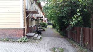 Trähusen i Port Arthur lockade paret Karl Österlund och Iiris Talvitie. Blommor, gröna buskar och träd vid ingången till arbetarträhus en sensommardag.