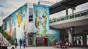 Suuri, värikäs, fantasiamainen seinämaalaus Myyrmäen juna-aseman ulkoseinissä, sinivihreitä hahmoja. 