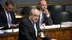 Kaj Turunen, fotograferad medan han talar under en session i riksdagen. I bakgrunden sitter Turunens nya partiledare Petteri Orpo (till vänster), och hans tidigare partiledare Sampo Terho (till höger). Riksdagsledamot Kaj Turunen talar, Petteri Orpo och Sampo Terho i bakgrunden.