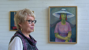 Åsa Hellman framför porträttet Flicka i vid hatt (Karin Hellman 1950, olja på papp). Kvinna står framför tavla av kvinna med grön hy.