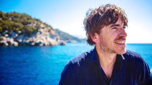 Simon Reeve Turkissa. Mies etualalla, hymyilee. Takana näkyy sininen meri ja kallioinen saari.