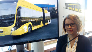 Ruut Haapamäki. Ruut Haapamäki står vid bilder av en gul superbuss och en gul spårvagn.