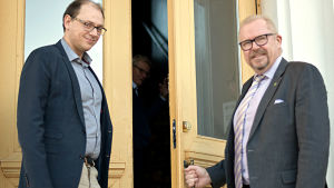 Styrelseordförande Anders Walls (till v.) och stadsdirektör Ragnar Lundqvist tror att Raseborg kommer på fötter igen. Två män i skjorta och kavaj, ena med slips, står på trappan till en gul stenbyggnad. Ena mannen håller dörren öppen för den andra. De ler och är på väg att stiga in i byggnaden.