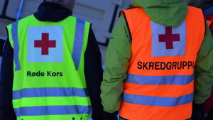 Två personer står med ryggen mot kameran. Den ena har en gul Röda Korset-väst. Den andra en röd väst där det står skredgruppen.