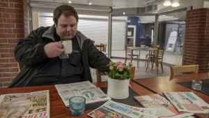 Taxichauffören Tor-Erik Stolpe sitter vid ett bord inne i en grillkiosk och dricker kaffe medan han läser tidningen.