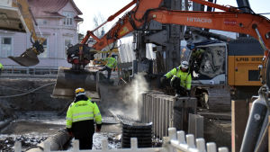 Arbetet pågår för fullt. En grävmaskin arbetar och några byggnadsarbetare övervakar.