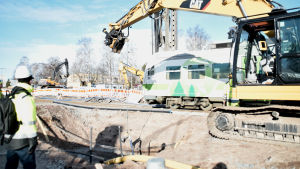 En rälsbuss passerar byggplatsen. Rälsbussen passerar en grävmaskin vid en grop.
