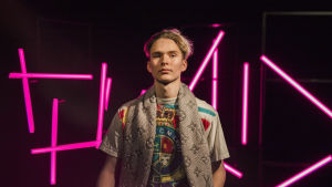Välillä omaisuudella flexaaminen on Iben mielestä coolia, toisinaan taas "ihan hirveeä". Vaaleahiuksinen nuori mies katsoo kameraan pinkkien neonvalotolppien edessä seisten, kuva rajattu vyötäröstä alaspäin.
