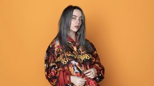 Laulaja Billie Eilish seisoo oranssia taustaa vasten.