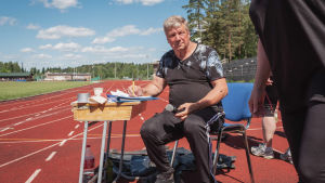 Jorma Rajakannas järjestää, kuuluttaa, kannustaa kaikkia osallistumaan ja heittää itsekin liitosaapasta. Mies istuu ulkona urheilukentällä pulpetti edessään ja mikrofoni ja kynä kädessään.