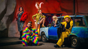 Viisi naista poseeraa auton ympärillä värikkäissä vaatteissa.
