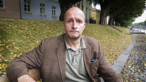 Christer Lindholm, en man utan hår sitter på en bänk vid Aura ås strand.