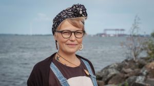 Vaaleatukkainen nainen silmälasit ja musta huivi päässä, taustalla merta