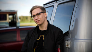 William i svart bomberjacka och en bandtshirt (elektrobandet Justice) logo framför bakdörrarna till en paketbil.