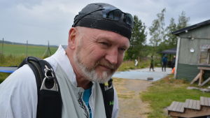 Pauli Mäki-Lohiluoma. Äldre man med skägg iklädd vit jacka står utanför en grön träbyggnad.