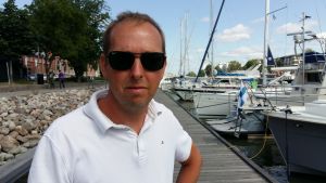 Kenneth Blomster. Kenneth Blomster, en man med solglasögon och vit pikéskjorta, står på en brygga med sgelebåtar.