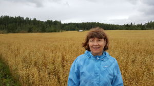 De torvåkrar vi har bör vi bevara och sköta om, säger Helena Äijö från täckdikningsföreningen. Helena Äijö står i förgrunden iklädd en blå vindjacka. Bakom henne ses sädesslag växa på en stor åker. Längst i bakgrunden skog och en grå, mulen himmel.