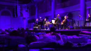 Sleep på Barbican i London år 2017. Människor ligger och sover på sängar framför en orkesterscen med en klassisk orkester.