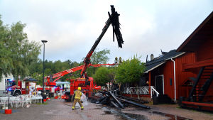 Morgonen efter branden jobbade räddningsverket fortfarande på. De måste riva taket för att kunna släcka elden. En brandbil med lyftkran lyfter trädbalkar från en röd bodas tak.