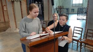 Sofia Liljeström visar för Mirjam Lillhannus och Iina Laakso vilka stycken som ska läsas ur Bibeln. Mirjam Lillhannus, en flicka med hästsvans och grå tröja, står vid en pulpet i Åbo Domkyrka. Pastor Sofia Liljeström visar vilket stycke som ska läsas ur Bibeln i samband med en högmässa.