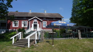 Rumarstrand är till salu. En röd stor stuga med vita knutar. På gräsmattan framför finns kafébord och kaféstolar.