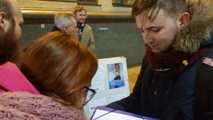 Jevgenij Karaulov samlar namn av förbipasserande. En ung man i en blå jacka med pälsliknande krage och en röd halsduk tittar ner på ett papper och framför honom står två personer med bakhuvudena mot kameran framåtlutade mot papperet den unga mannen håller i.